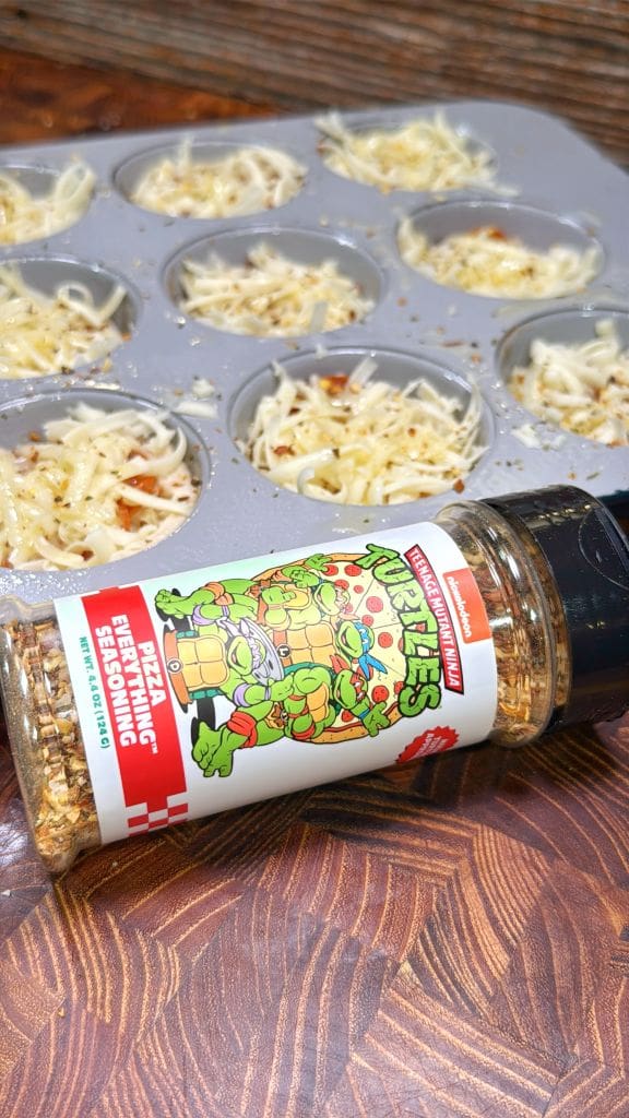 A seasoning bottle with an illustration of cartoon turtles rests on a wooden surface. Behind it, a muffin tin is filled with cheese and toppings, ready for baking.