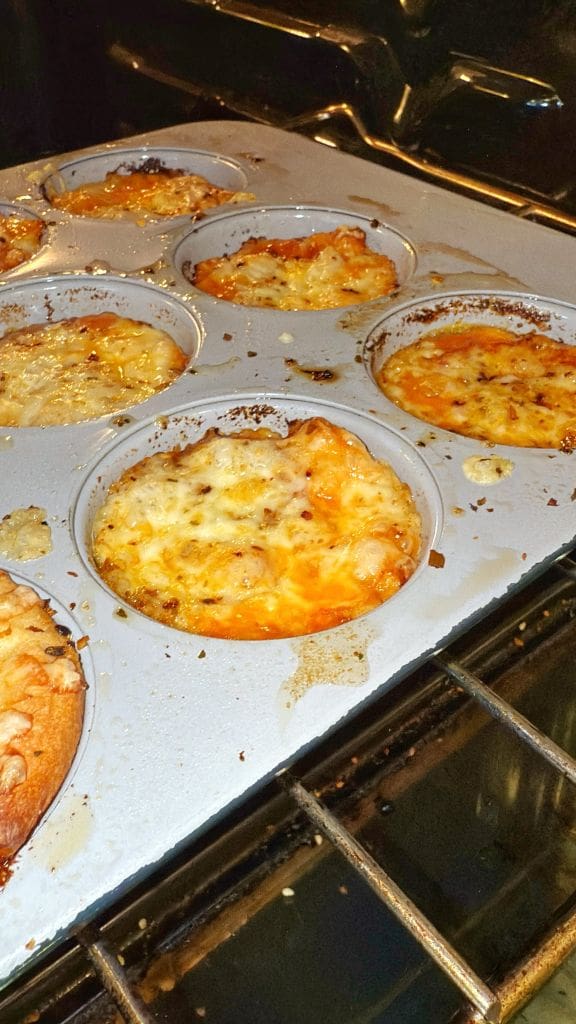 Muffin tin filled with baked egg and cheese muffins in a metal tray inside an oven. The muffins are golden brown with melted cheese on top. Some cheese has spilled over the edges of the tin.