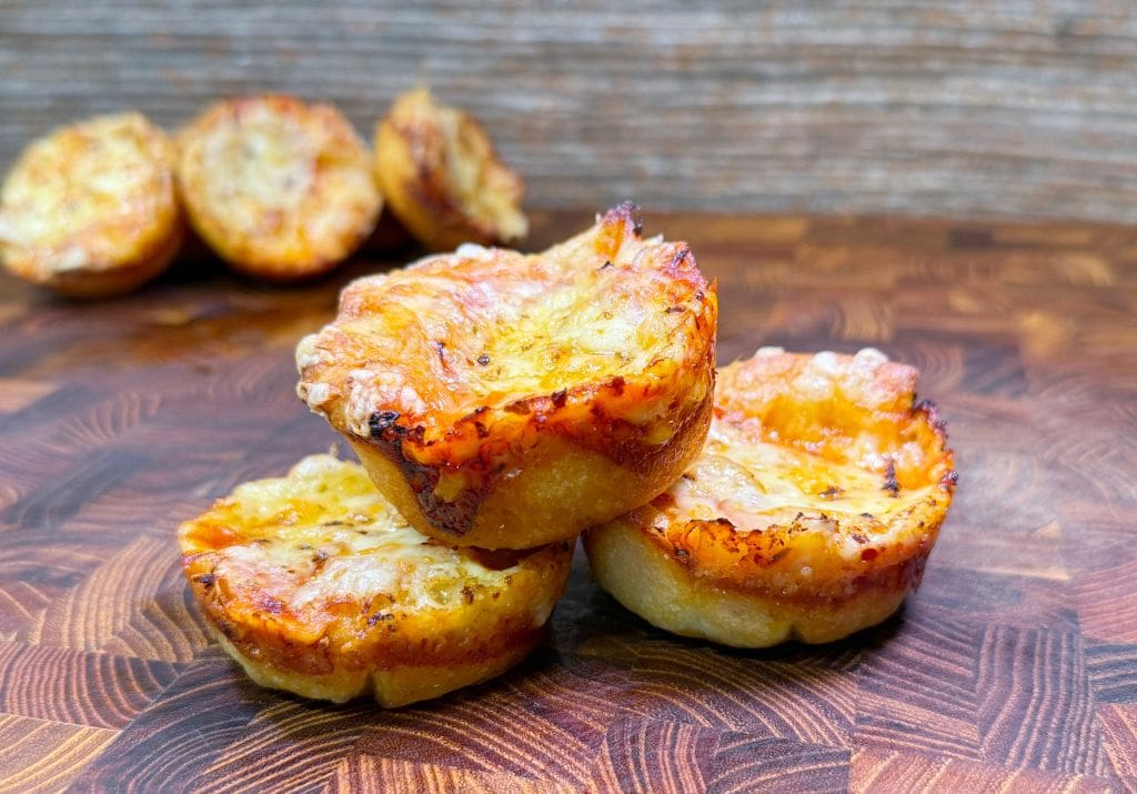 Three baked mini muffin pizzas with melted cheese and slightly crisp edges are stacked on a wooden surface. More mini pizzas are visible in the blurred background. Image is of a crazy puffs recipe