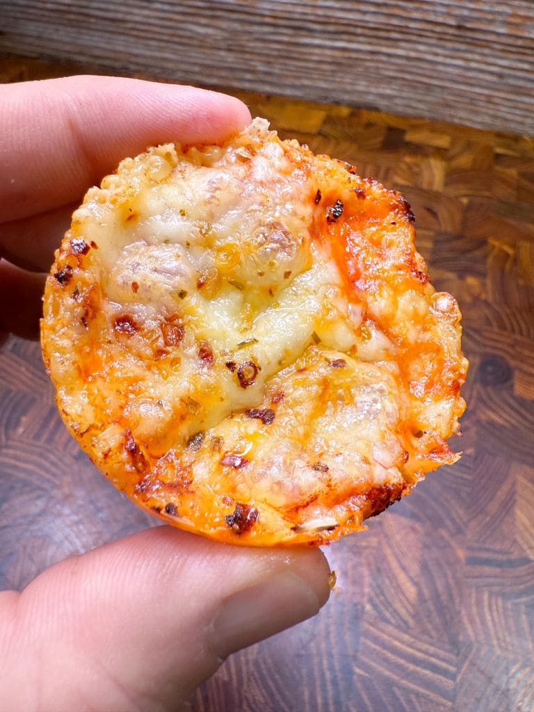A hand holding a small pizza muffin with melted cheese and a crispy, golden-brown topping. The wooden background adds a rustic touch to the image.