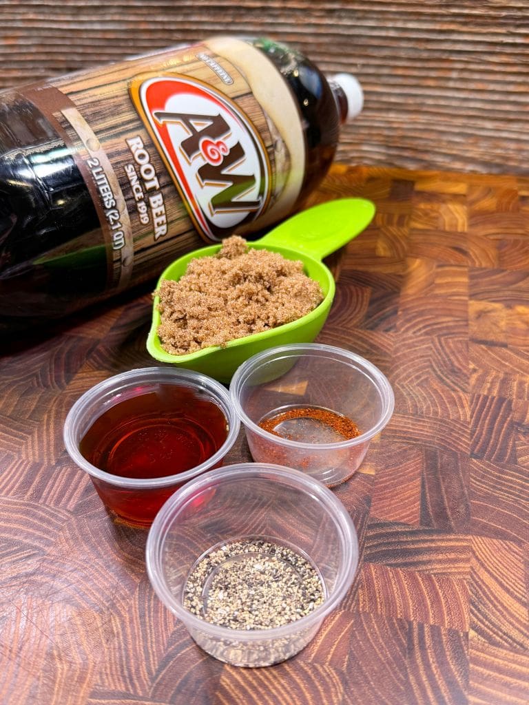 A bottle of A&W Root Beer lies on its side next to a green measuring cup filled with brown sugar. In front are small containers with red liquid, a spice blend, black pepper, and another dark spice on a wooden surface.