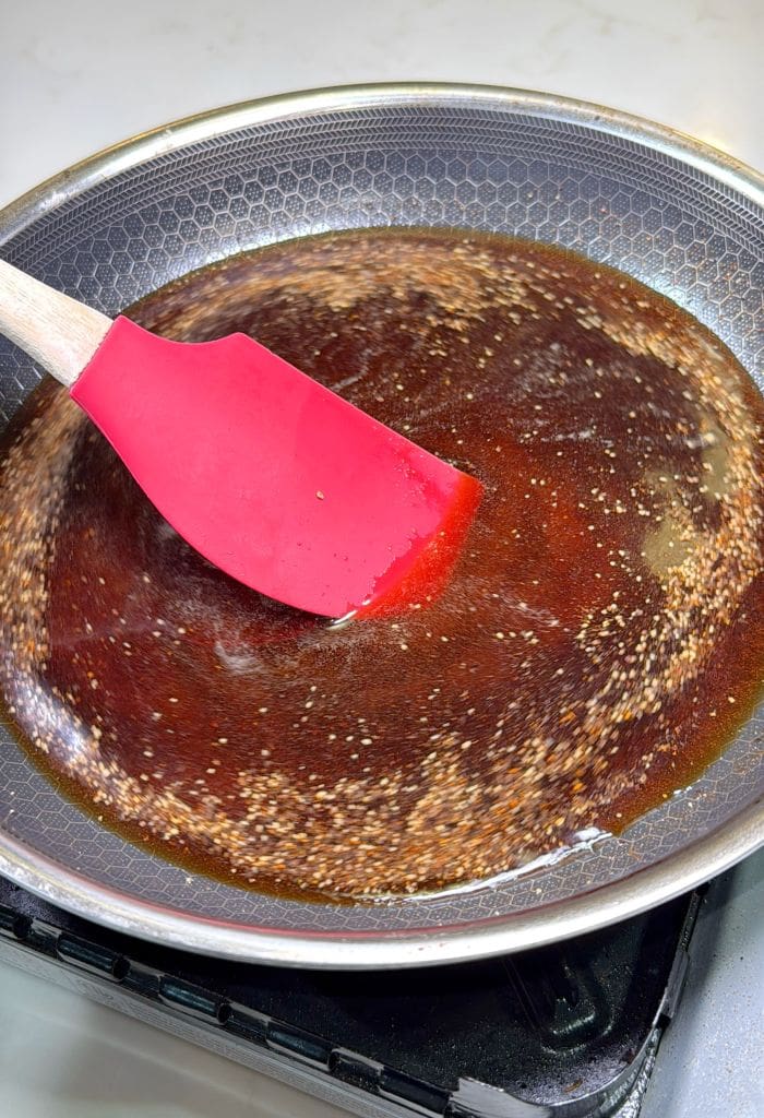 A red spatula stirs a dark brown sauce with floating sesame seeds in a nonstick frying pan on a stovetop.