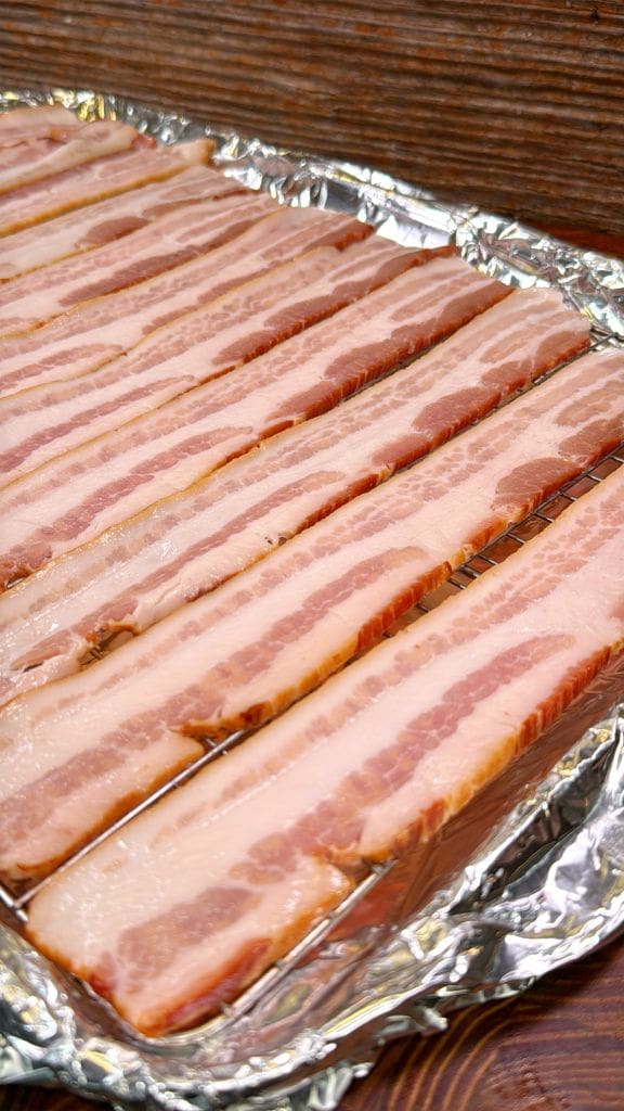 Strips of uncooked bacon lined up on a foil-covered baking sheet, ready for cooking. The bacon shows distinct layers of pink meat and white fat.
