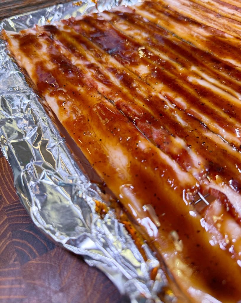 Close-up of raw ribs covered in a rich barbecue sauce, placed on a foil-lined surface. The ribs appear well-seasoned and ready for cooking. The sauce glistens under the light, enhancing the texture and color of the meat.