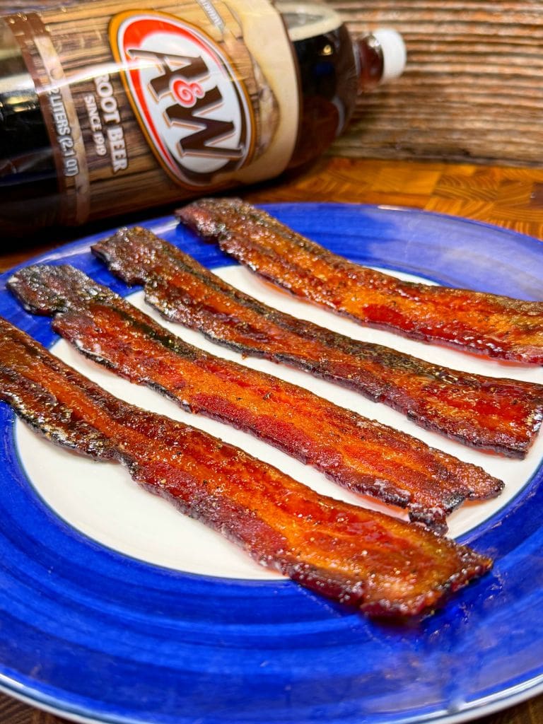 Crispy candied bacon strips arranged on a vibrant blue and white plate, with an A&W root beer bottle lying nearby on a wooden surface. The bacon appears glazed and caramelized, contrasting with the rustic background.