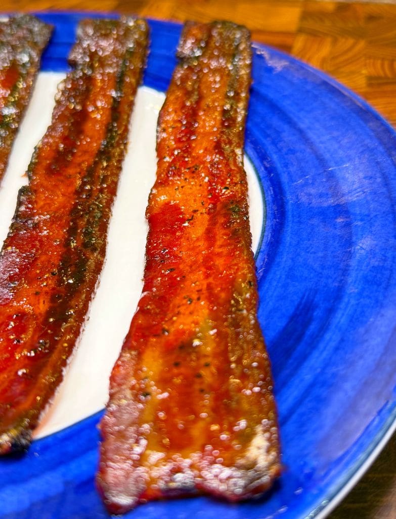 Close-up of three crispy, caramelized bacon strips on a bright blue plate, showcasing a glossy, slightly glazed surface with a hint of pepper and spices. The bacon appears freshly cooked and appetizing.