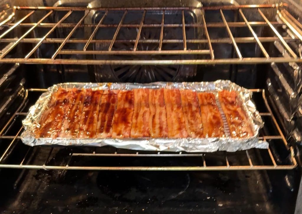 Strips of root beer candied bacon lined up on a foil-covered baking tray in an oven, with the bacon glistening as it cooks.