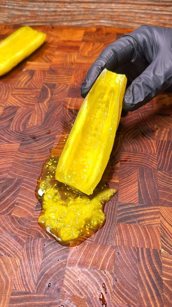 A gloved hand holds a halved pickle on a wooden cutting board. Pickle juice and seeds are visible on the board. Another halved pickle lies nearby.