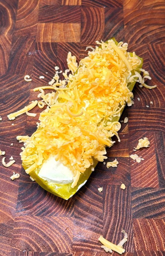 A large pickle sliced in half, filled with cream cheese, and topped with shredded cheddar cheese sits on a wooden cutting board. Shredded cheese is sprinkled around it.