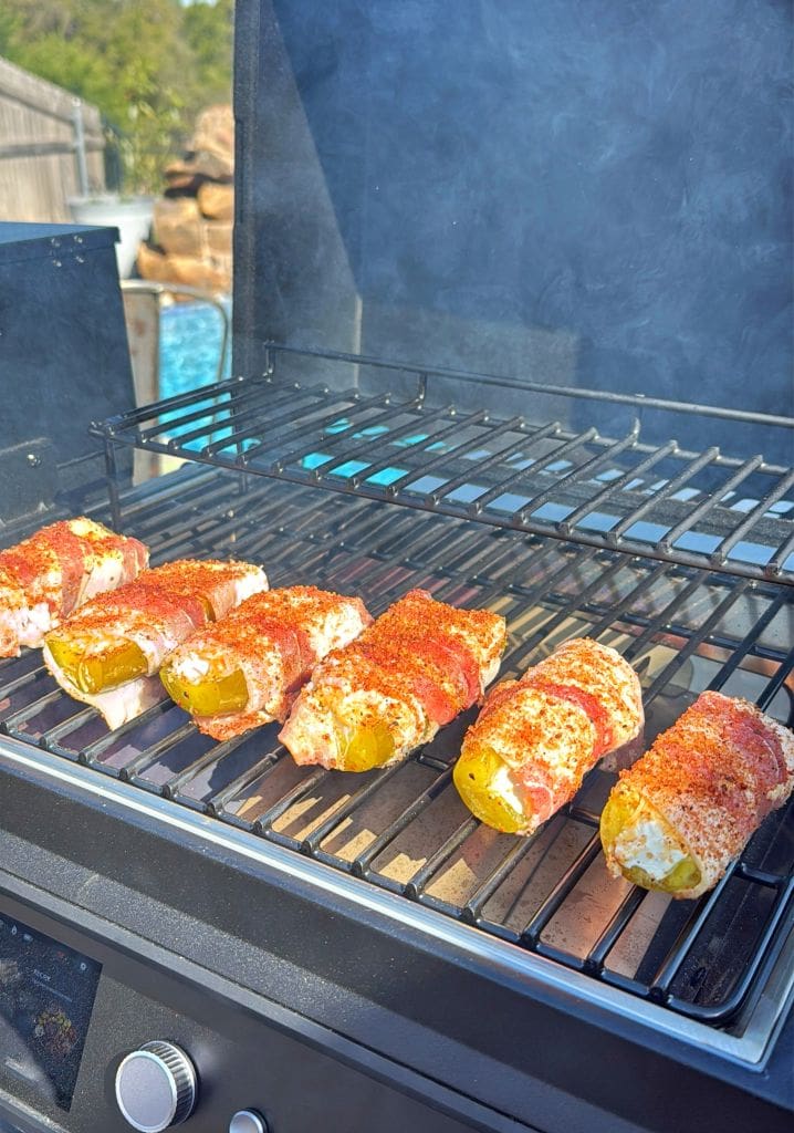 Chicken breasts wrapped in bacon and seasoned with spices are cooking on a grill. The grill is outdoors near a pool, and smoke is rising from the sizzling food.