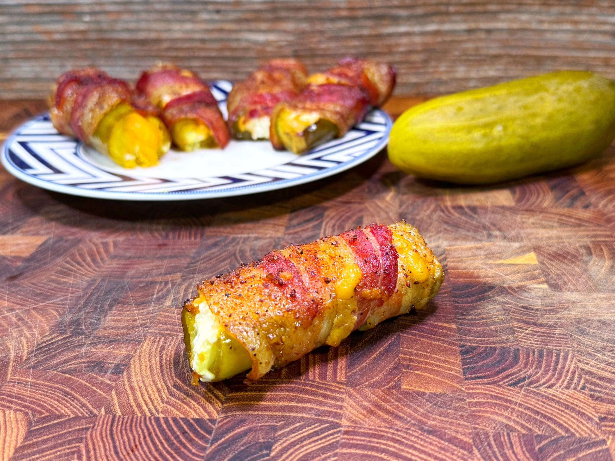 A bacon-wrapped, cheese-stuffed pickle is in the foreground on a wooden cutting board. In the background, a plate holds more stuffed pickles, and a whole pickle lies beside the plate. The focus is on the detailed texture of the stuffed pickle.