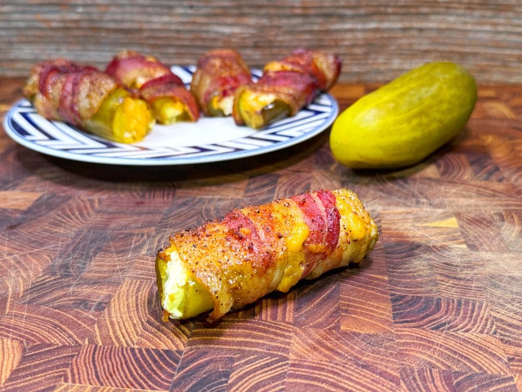 Bacon-wrapped stuffed jalapeños on a wooden surface, with one in the foreground. A plate with more jalapeños and a whole pickle are in the background. The jalapeños appear to be stuffed and baked.