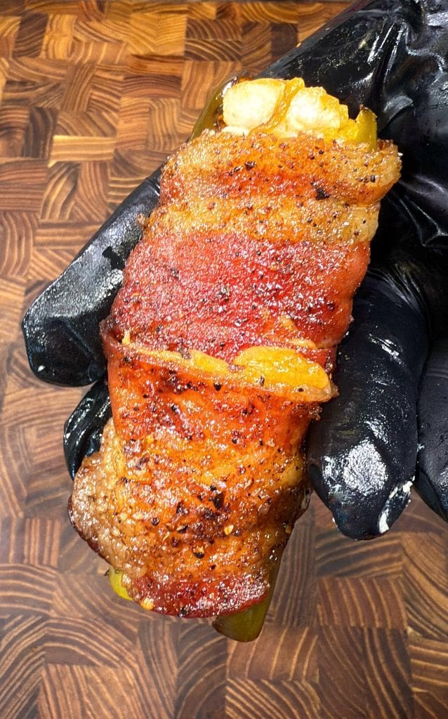 A gloved hand holding a jalapeño stuffed with cheese, wrapped in crispy bacon, seasoned with pepper and spices. The background is a wooden cutting board.