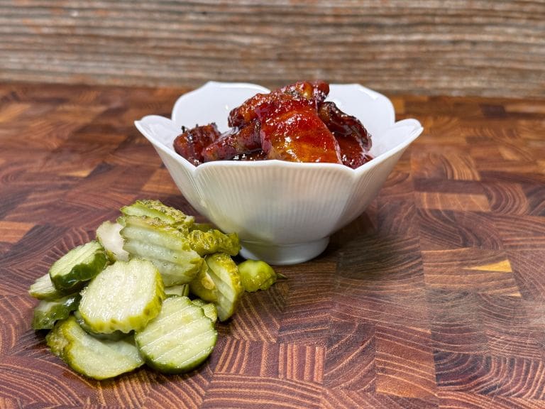 A white bowl filled with glazed, grilled meat is on a wooden surface. In the foreground, there are slices of pickles arranged neatly. The background features a textured wood pattern.
