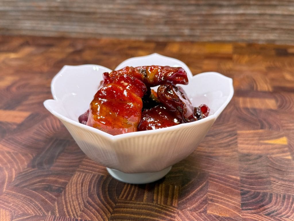 A shiny, dark red dish of glazed shrimp served in a decorative white bowl on a wooden table with a geometric pattern.