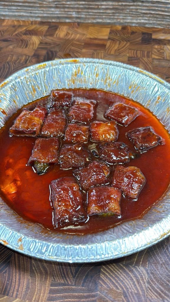 A disposable foil tray filled with glazed, sauce-covered chunks of meat, resting on a wooden surface. The sauce is thick and red, coating the meat pieces evenly.