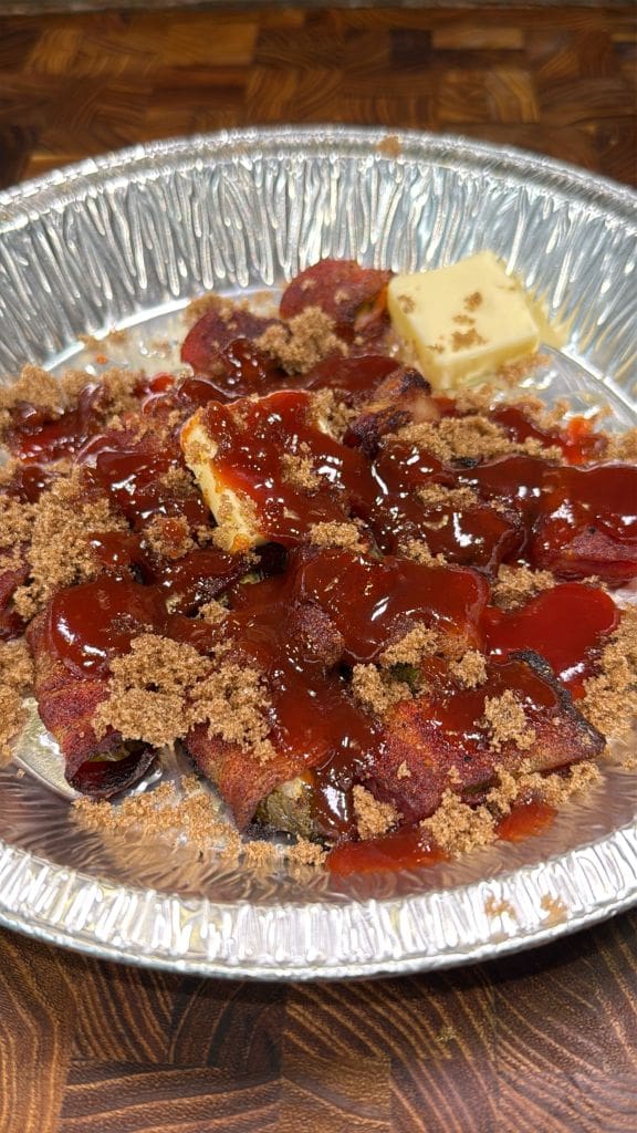 A silver foil tray filled with pickle burnt ends wrapped in bacon strips topped with brown sugar and drizzled with reddish sauce. A small piece of butter is visible on the side. The tray sits on a wooden surface.