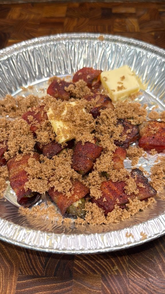 A foil tray holds bacon-wrapped jalapeños topped with brown sugar and two pats of butter, placed on a wooden surface.