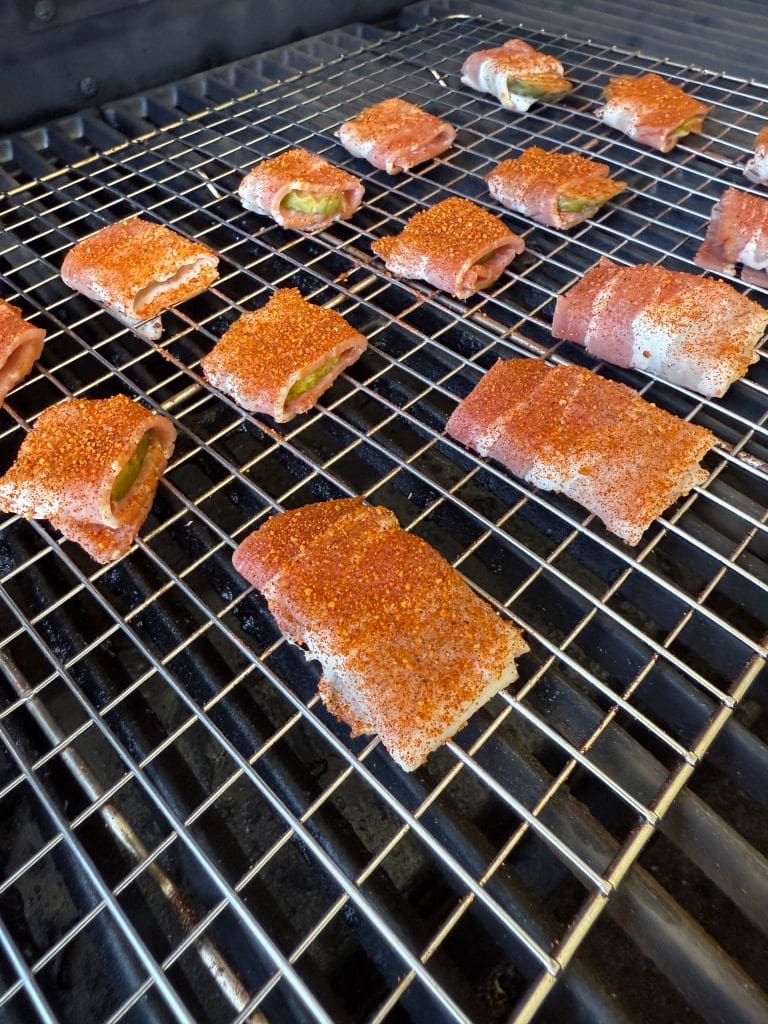Jalapeño poppers wrapped in bacon and coated with seasoning are lined up on a grill, cooking. The grills black grates contrast with the sizzling appetizer, highlighting the pops of green from the jalapeños and the reddish-brown seasoning.