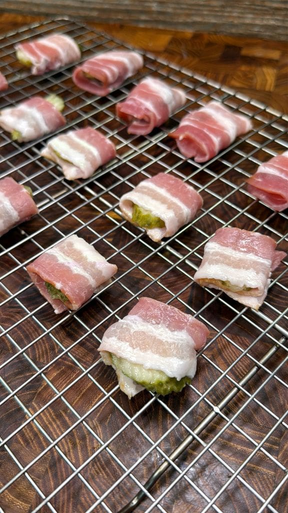 Slices of bacon wrapped around pieces of bread with green filling, placed on a metal wire rack. The preparation is on a wooden surface, ready for cooking.