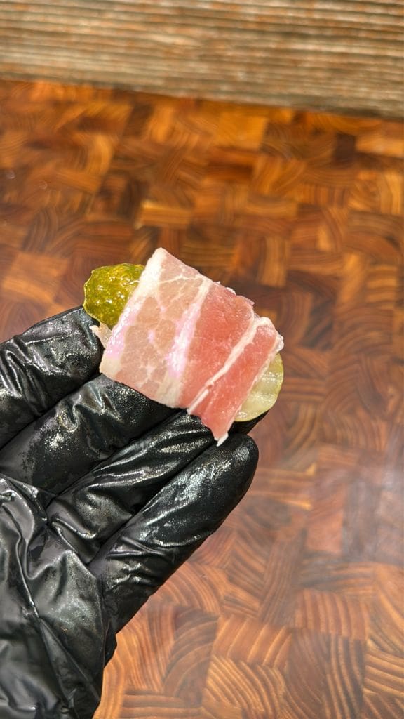 A hand in a black glove holds a small piece of food with a slice of pink meat wrapped around something green, likely a pickle. The background shows a wooden surface with a geometric pattern.