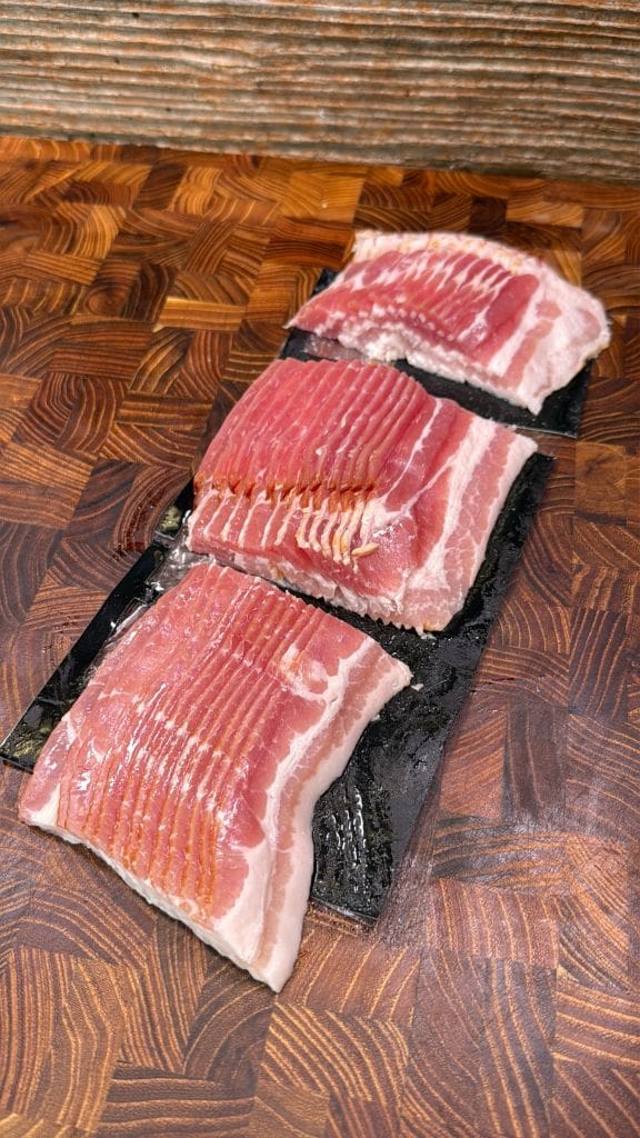 Three stacks of thinly sliced raw bacon are arranged on a dark, glossy surface. The bacon showcases a striped pattern of pink meat and white fat. The background features a textured, brown wooden cutting board with a rich grain pattern.