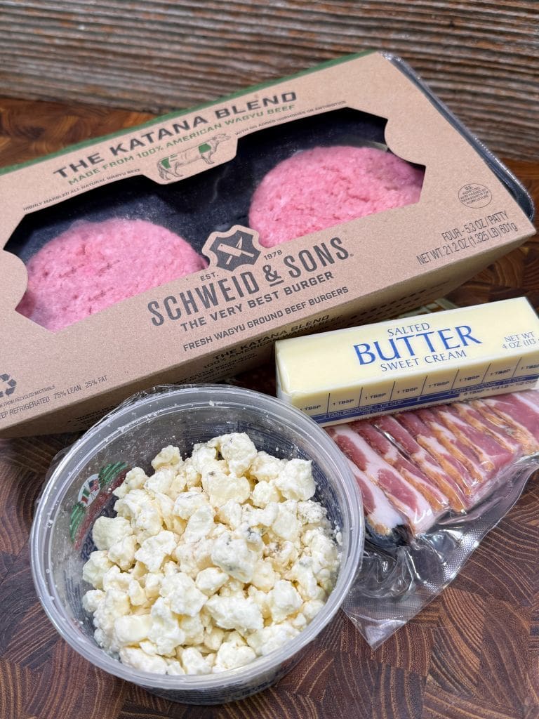A package of Schweid & Sons ground beef patties, a stick of salted sweet cream butter, a pack of sliced bacon, and a container of crumbled cheese on a wooden surface.