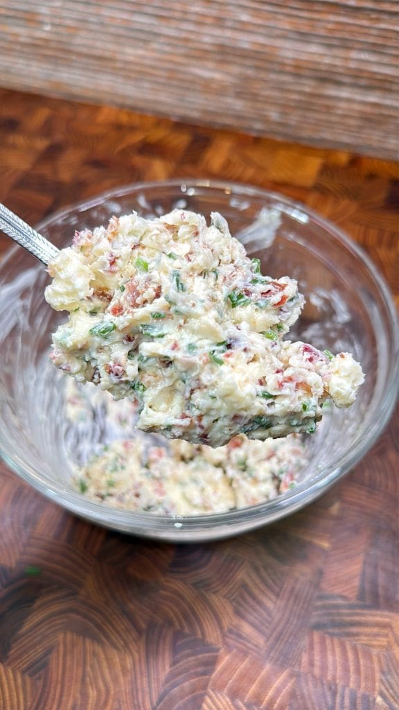 A spoon lifts a mixture of finely chopped herbs, bacon bits, and a creamy base from a glass bowl, set on a wooden surface.