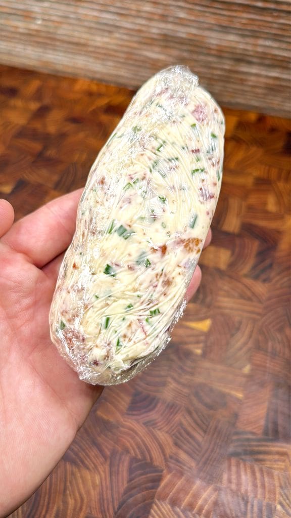 A hand holds a log of compound butter mixed with herbs and possibly bits of dried meat, tightly wrapped in plastic wrap, over a patterned wooden surface.