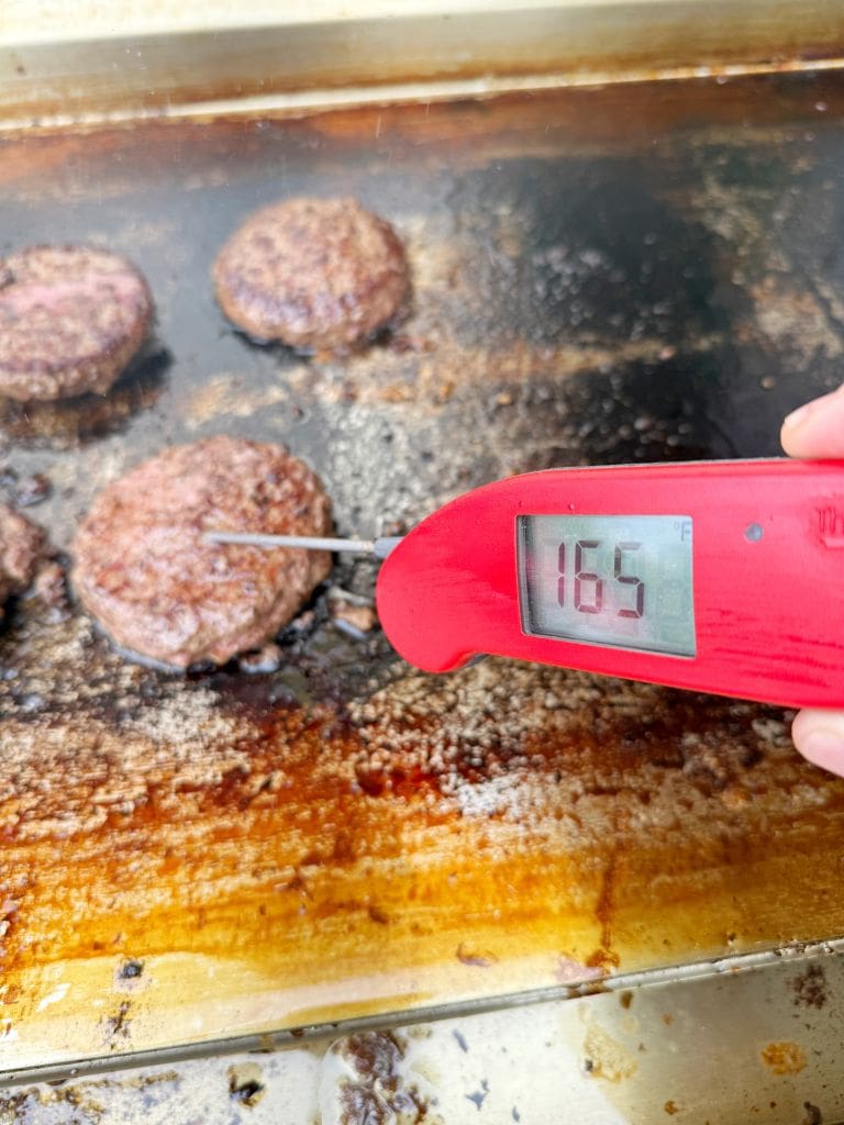 A hand holds a red digital meat thermometer inserted into a hamburger patty on a grill. The thermometer displays a temperature of 165°F. Several other patties are also cooking on the greasy grill surface.