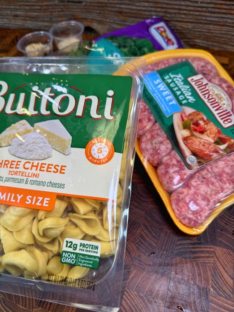 A package of Buitoni three cheese tortellini, a pack of Johnsonville sweet Italian sausage, and a bag of frozen spinach are arranged on a wooden surface.