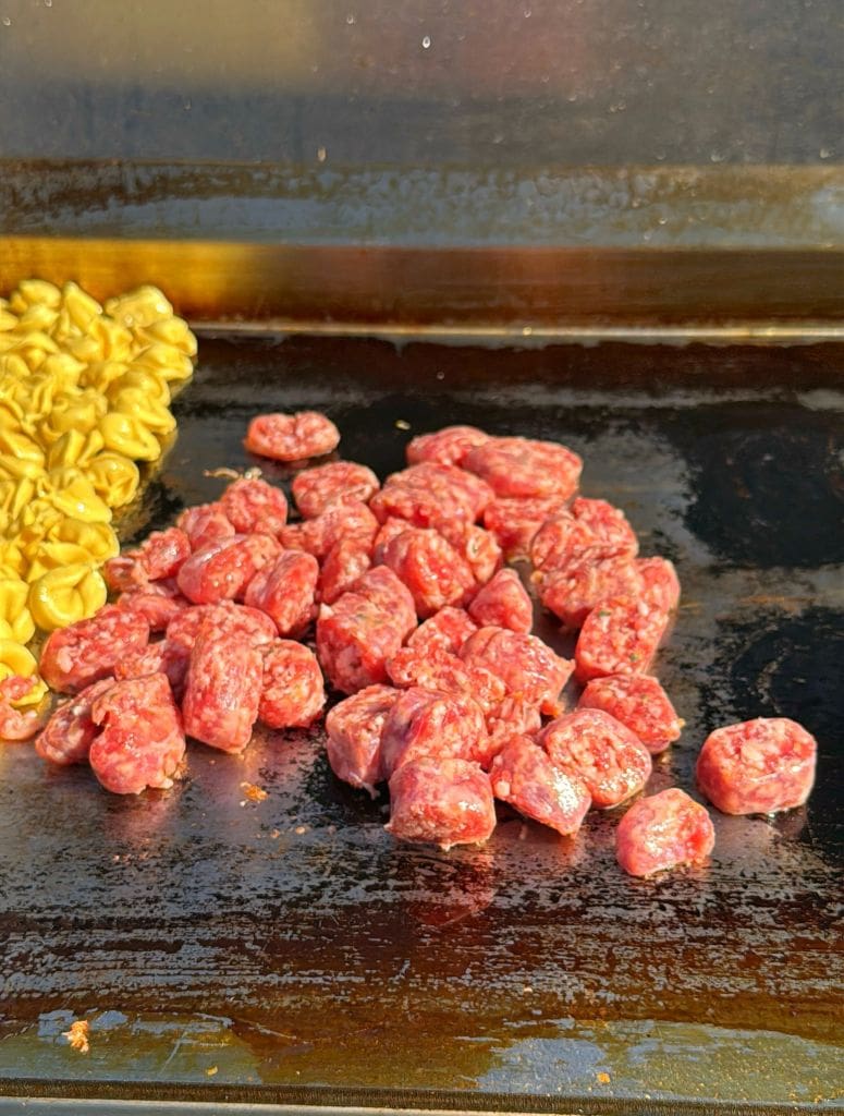 Chunks of raw sausage and cooked tortellini pasta are sizzling on a flat top grill or griddle surface. The sausage is uncooked and red, while the tortellini is yellow and appears cooked.