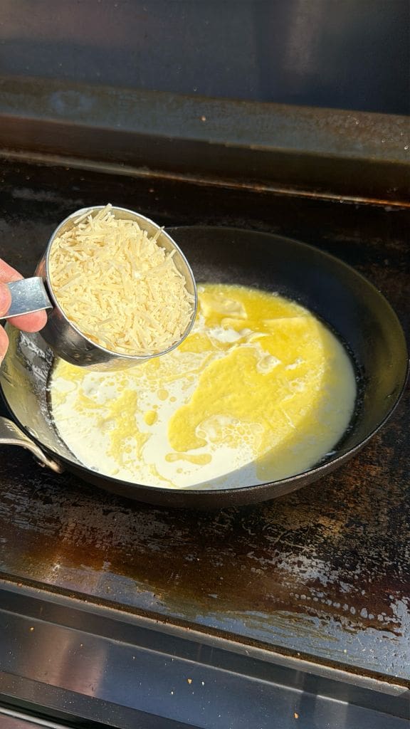 A hand holds a measuring cup of uncooked rice over a skillet with melted butter and cream, ready to be added and cooked on a stovetop.