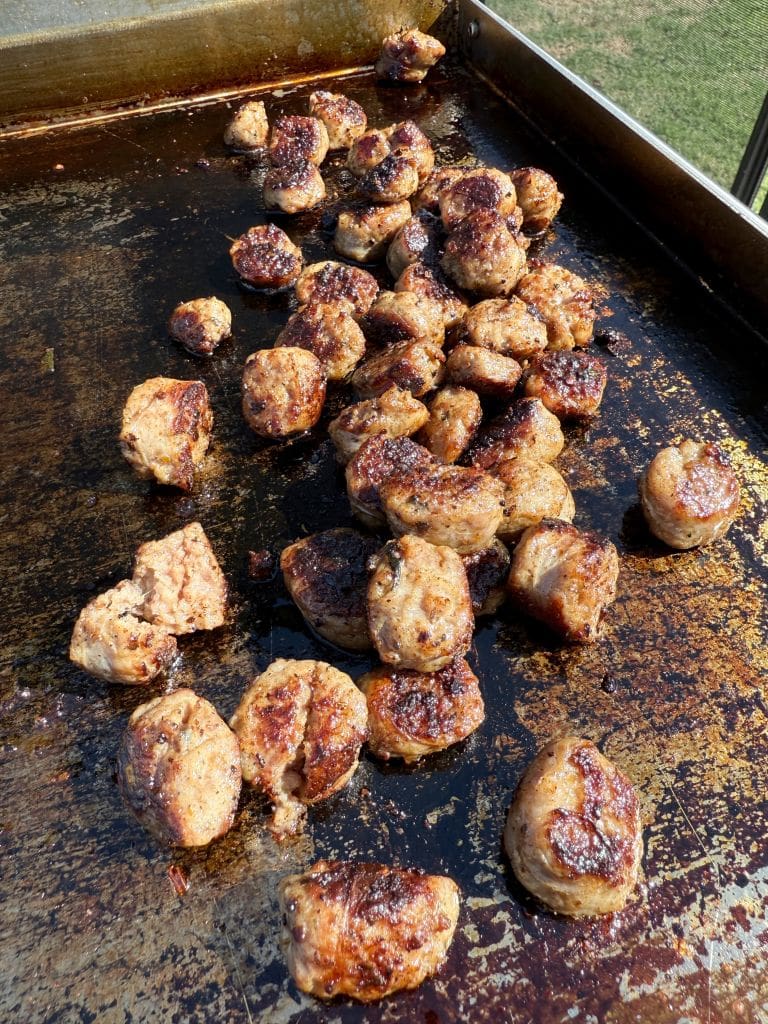 Chunks of grilled meat with a browned, crispy exterior are sizzling on a flat-top grill outside, with a patch of green grass visible in the background.