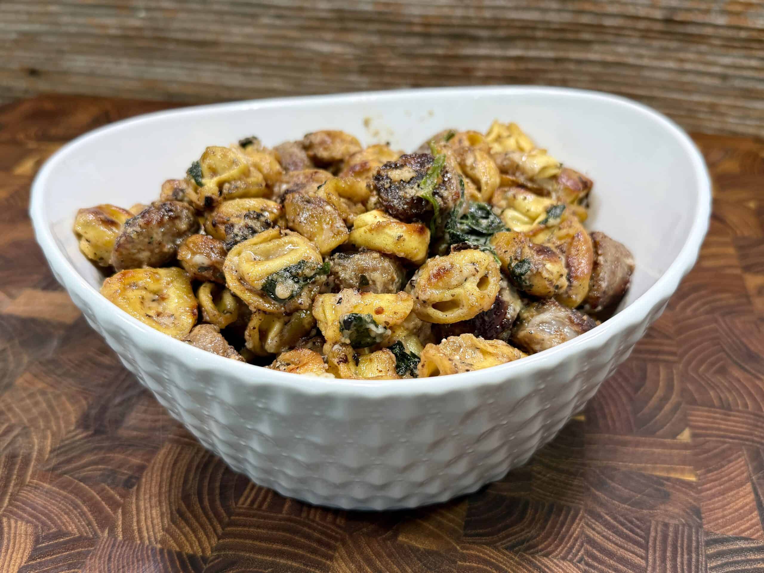 A white textured bowl filled with cooked tortellini pasta, sausage slices, and leafy greens sits on a wooden surface with a rustic wood background.