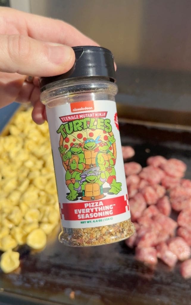 A hand holds a Teenage Mutant Ninja Turtles Pizza Everything Seasoning bottle over a grill with pasta and sausage cooking in the background.