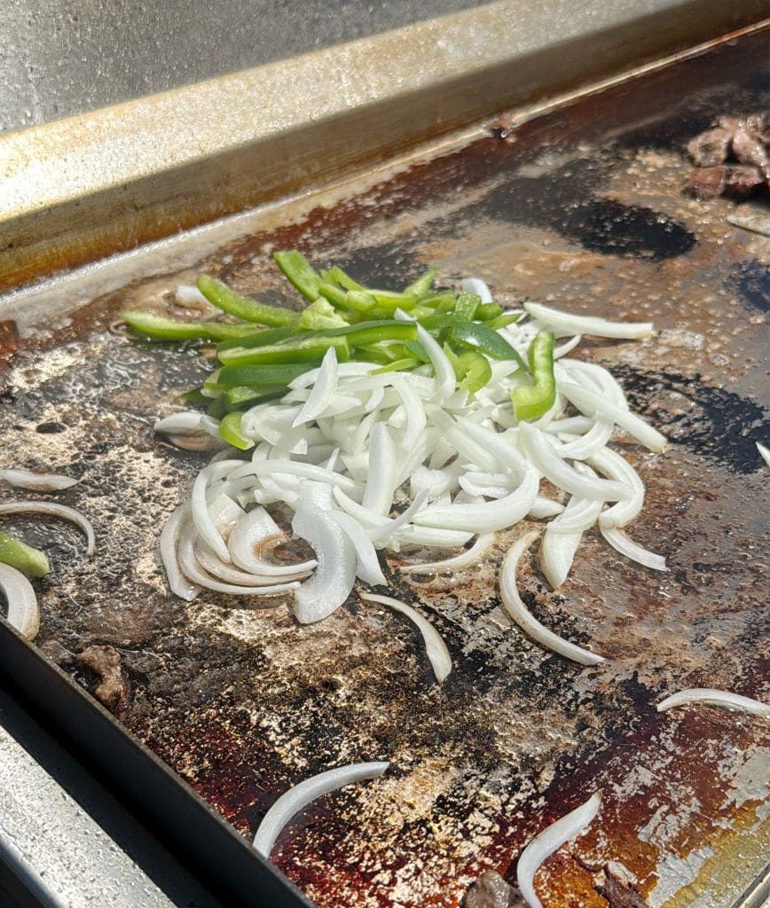 Sliced onions and green bell peppers are cooking on a flat top grill, with some browning and residue visible on the hot surface under bright sunlight.