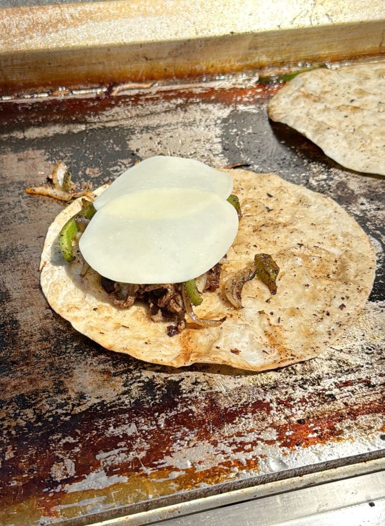 A tortilla filled with grilled beef, onions, and green peppers, topped with two slices of white cheese, cooking on a griddle beside another plain tortilla.