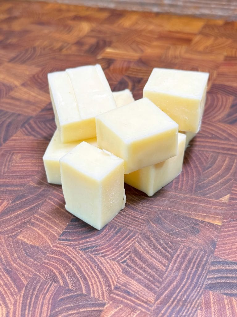 Several small cubes of pale yellow cheese are stacked on a wooden cutting board with a patterned surface.