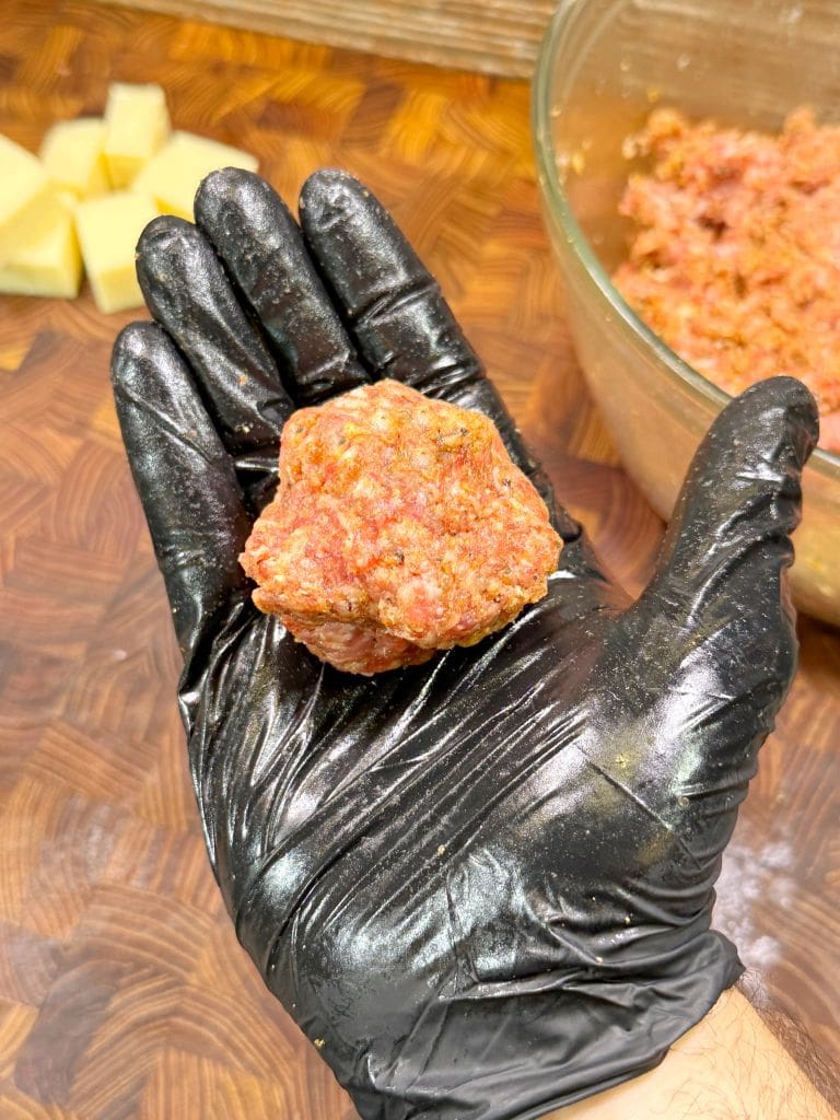 A hand wearing a black glove holds a raw meatball over a wooden surface. A glass bowl with more ground meat and some potato pieces are visible in the background.