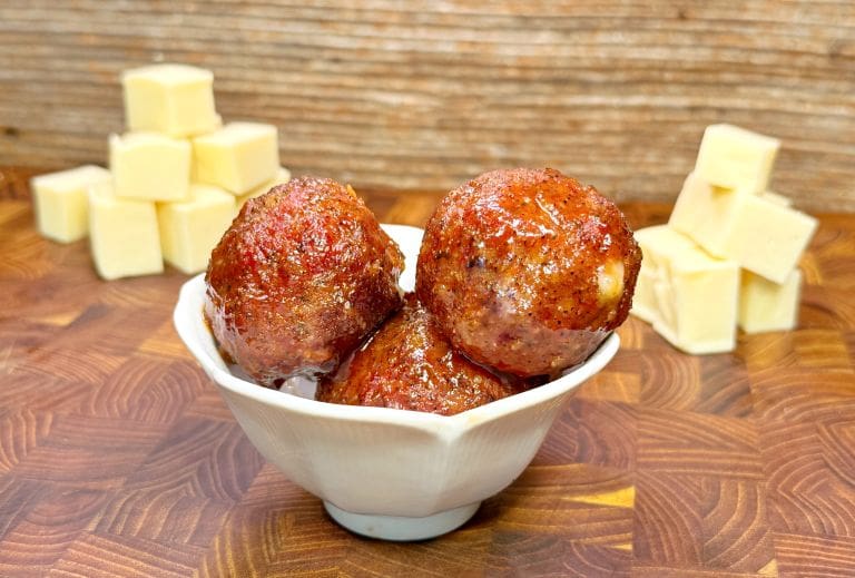 A white bowl holds three glazed meatballs, with cubes of cheese stacked in the background on a wooden surface.