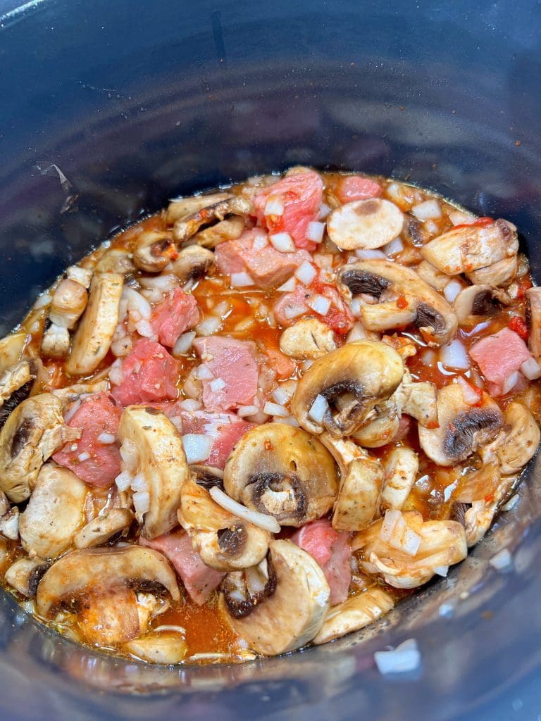 A close-up of a slow cooker filled with sliced mushrooms, diced tomatoes, chopped onions, and seasonings in a tomato-based sauce for this crockpot beef tips recipe.