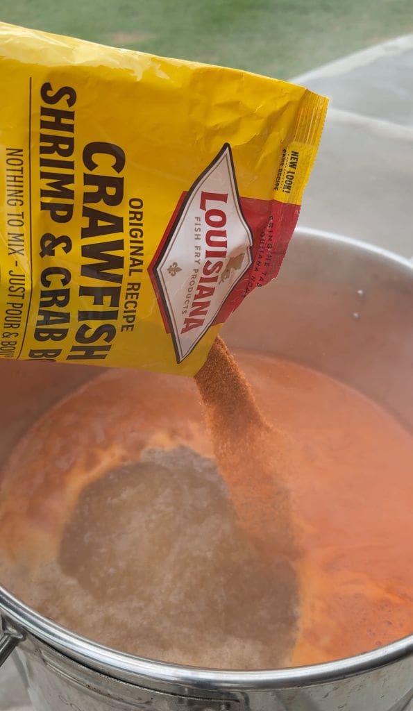 A hand pours Louisiana Crawfish, Shrimp & Crab Boil seasoning from a yellow packet into a steaming pot of boiling water.