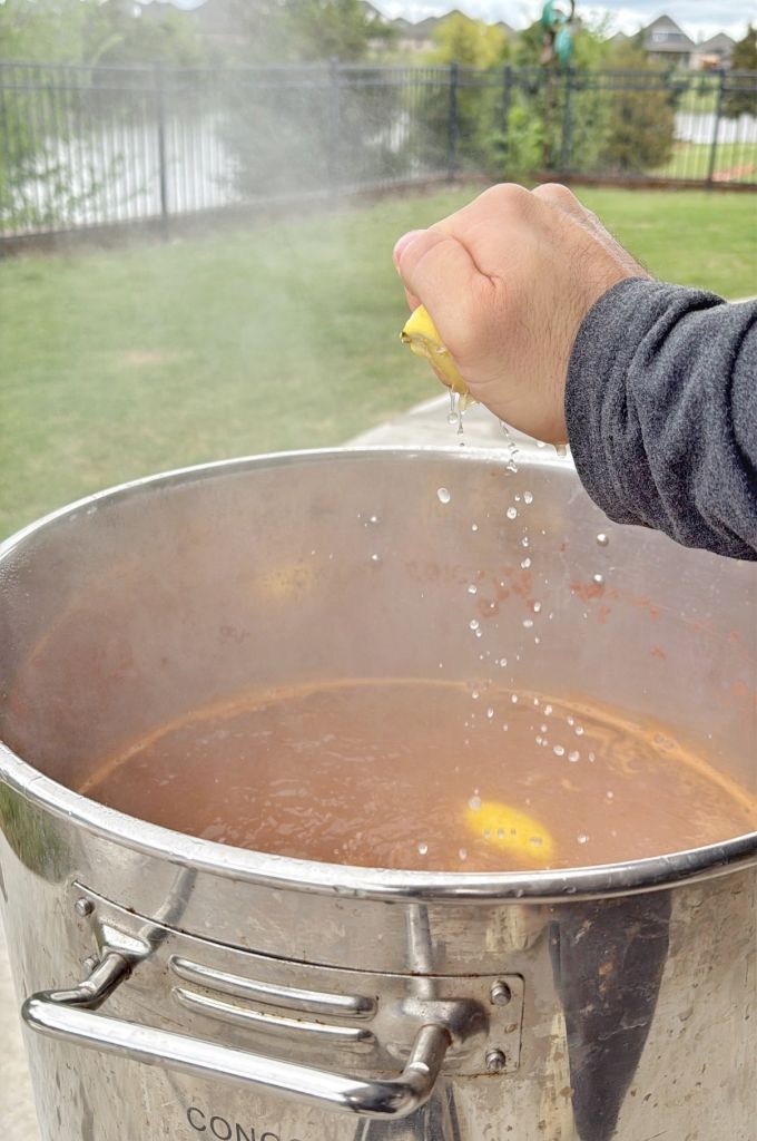 A person squeezes a lemon over a large steaming metal pot outdoors, letting juice and seeds fall into the simmering liquid. Trees, grass, and a fence are visible in the background.