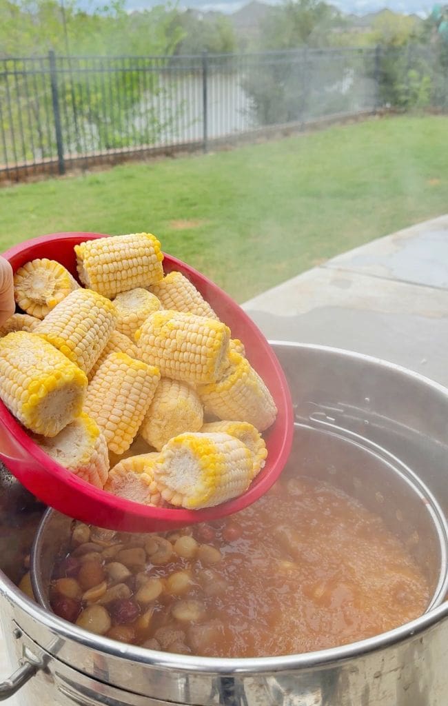 A hand holds a red bowl of corn on the cob pieces over a steaming outdoor pot filled with boiling sausage, potatoes, and other ingredients, ready to add the corn to the mix. A yard and fence are visible in the background.