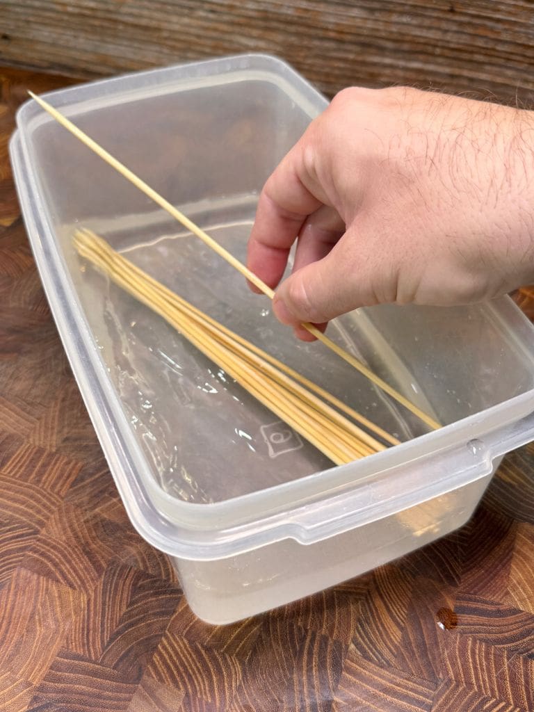 A hand places a wooden skewer into a clear plastic container filled with water, where several other skewers are already soaking.