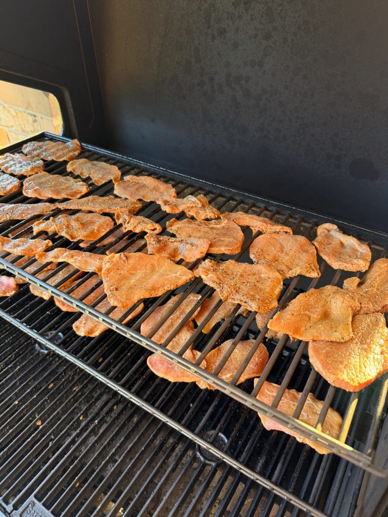 Thin slices of seasoned meat are arranged on the upper and lower racks of a black outdoor grill, cooking over the heat. Sunlight shines on the meat, highlighting its golden brown color.