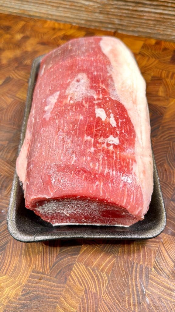 A large, raw cut of beef with a layer of fat on one side sits on a black tray on a patterned wood surface.