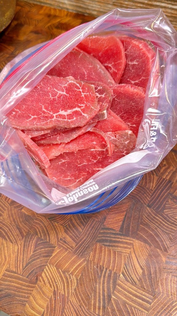 Raw slices of red beef placed inside a resealable plastic bag, resting on a wooden countertop with a distinctive geometric pattern.