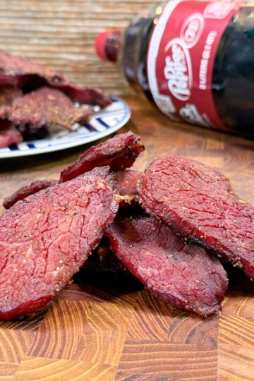 Slices of beef jerky stacked on a wooden surface with a plate of more jerky and a bottle of Dr Pepper soda in the background.